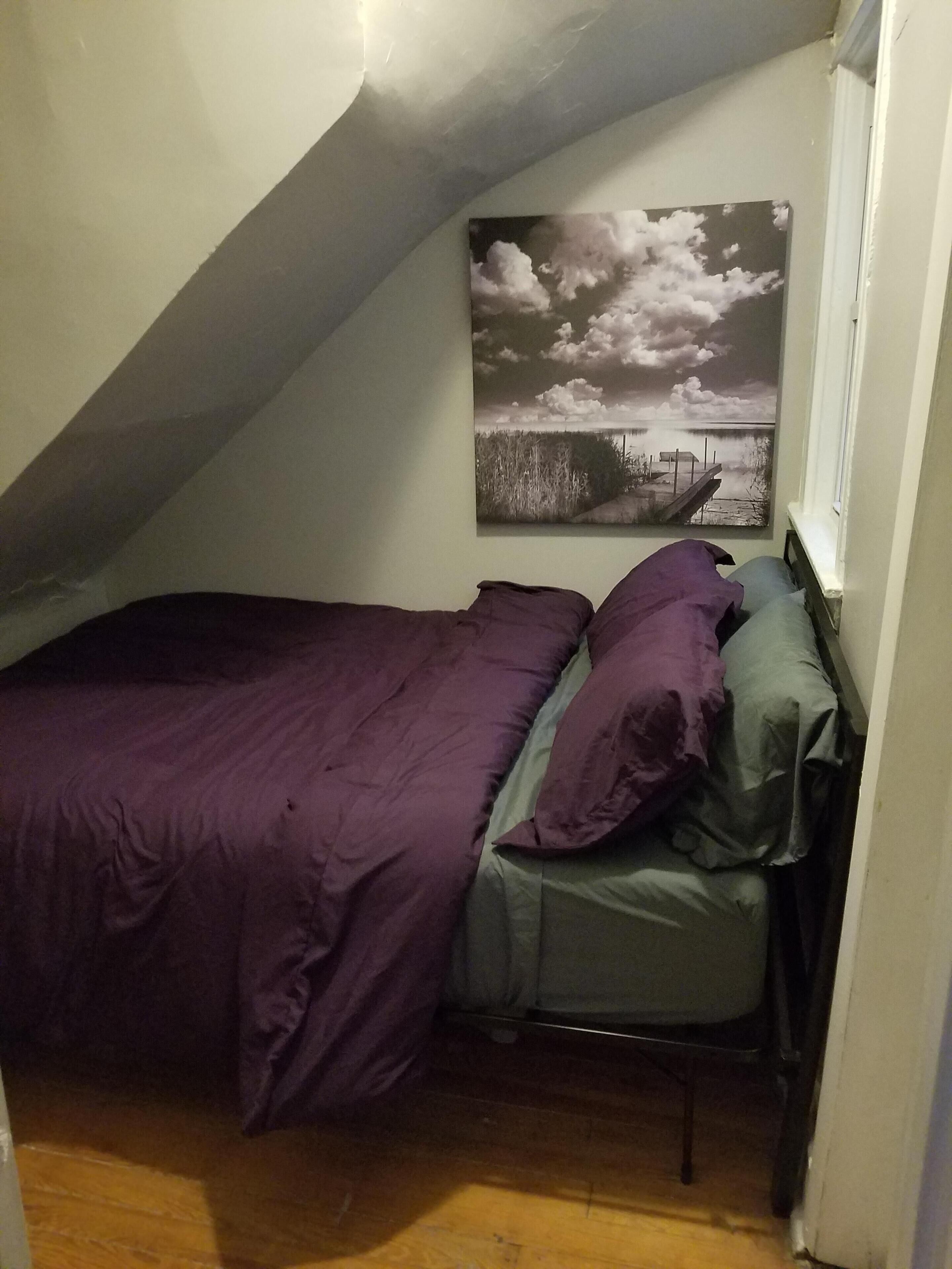 Une petite pièce à la lumière tamisée, avec un plafond en pente, un lit recouvert d’une literie violette, une seule chaise et un tableau accroché au mur représentant un ciel nuageux et de l’eau.