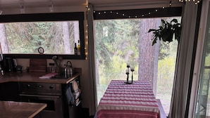 Una cocina acogedora con comedor, que incluye una mesa con mantel rojo y blanco, una planta colgante y vistas a un bosque a través de grandes ventanales.