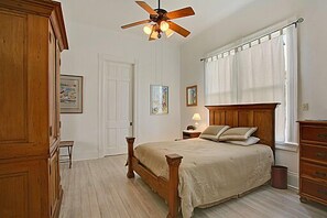 Un dormitorio con una cama de madera, un ventilador de techo, una mesita de noche con lámpara y una ventana con cortinas.