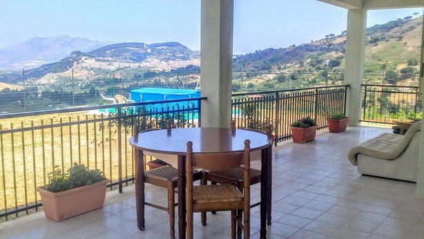 A balcony with a round table and chairs, potted plants, and a view of a coastal area with mountains in the background. - Nice place,organic garden,quiet,history of Italy,enchanting views Segesta (Calatafimi Segesta)