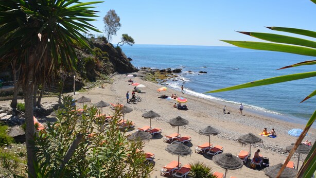 Une plage avec des parasols en chaume et des chaises longues, un rivage rocheux et une eau d’un bleu limpide.