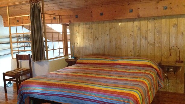 A wooden cabin room with a bed, a chair, a window with curtains, and a lamp. - Valtellina mon amour (Castionetto)