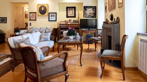 Un salon comprenant un canapé, des fauteuils, une table basse et une cheminée. Les murs sont ornés de tableaux encadrés et un téléviseur est posé sur un meuble.