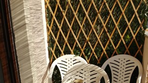 Ein kleiner Sitzbereich im Freien mit einem Tisch und zwei weißen Plastikstühlen. Der Tisch ist gedeckt mit einer Auswahl an Gebäck, zwei Tassen und einem Glas rotem Getränk.