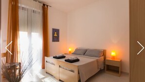 Ein Schlafzimmer mit Bett, Nachttischlampen, einem Nachttisch, einem Sessel, einem Fenster mit Vorhängen und einem Wandbild.