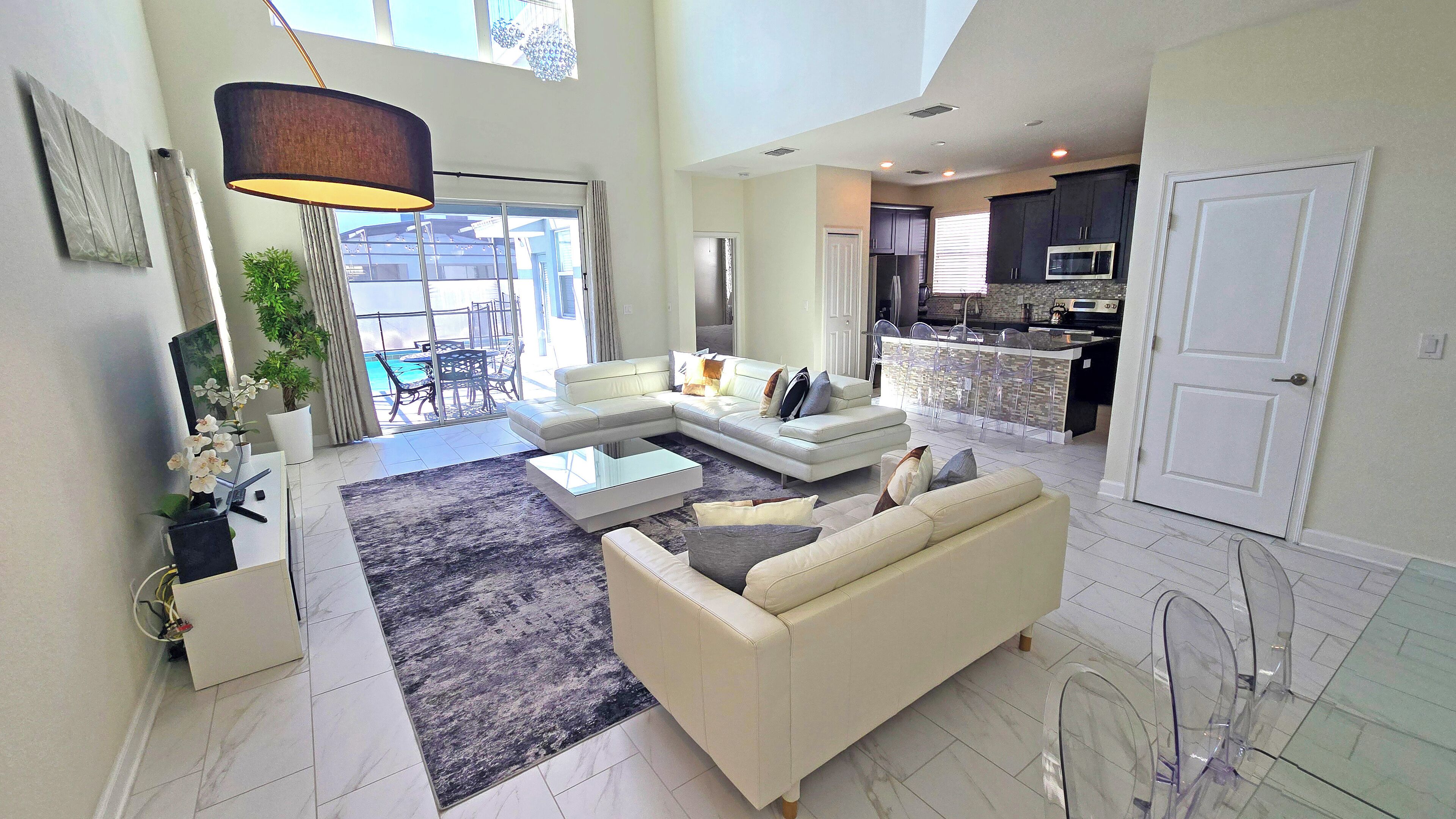 A spacious living area with a large L-shaped sofa, a coffee table, and a dining area with a glass table and chairs. There is a kitchen with dark cabinets and a white door.