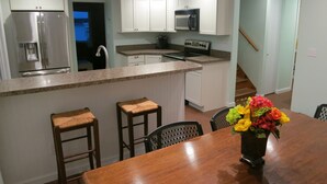 Una cocina moderna con armarios blancos, un refrigerador de acero inoxidable, un microondas integrado y una encimera de granito. Hay dos taburetes de bar de madera y una mesa de comedor de madera con un florero.