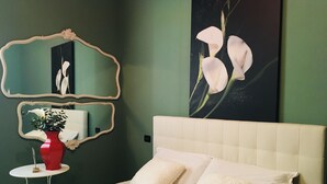 A bedroom with a bed, pillows, a red vase with a plant, a mirror, and a painting of flowers on the wall.