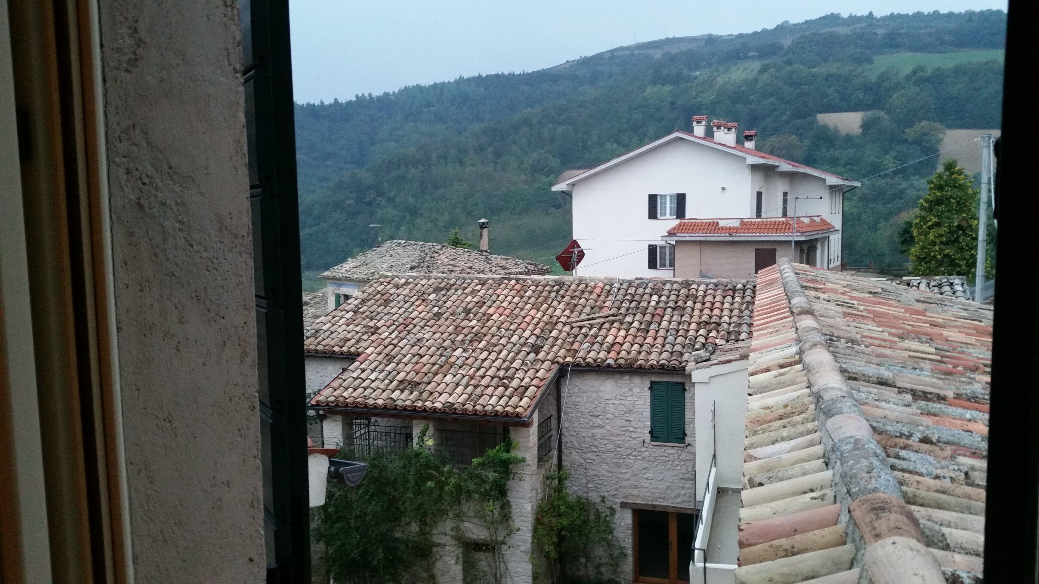 A view of a hillside village with traditional buildings, including a house with a red-tiled roof and a stone wall.