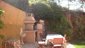 Une terrasse extérieure avec une table en pierre, des chaises en bois, un four en briques et une petite table recouverte d’une nappe à motifs.