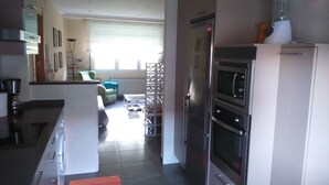 Eine moderne Küche mit Edelstahlgeräten, ein Essbereich mit einem grünen Stuhl und ein Blick auf das Wohnzimmer durch eine Glaswand.