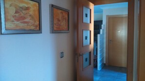 A hallway with framed artwork on the walls, a door leading to a room with a tiled floor, and a shelving unit in the background.
