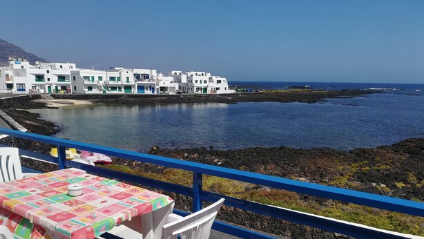A balcony with a table and chairs overlooking a coastal area with buildings and a body of water. - Odamar Órzola apartament (Órzola, Lanzarote)