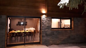 Un bâtiment en bois avec un espace repas visible à travers des portes vitrées, un mur en pierre et une fenêtre donnant sur l’intérieur.