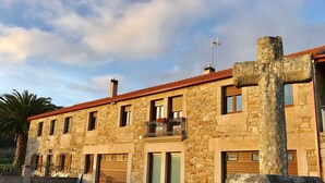 A stone building with multiple windows, a balcony, and a large stone cross in the foreground. - House in a natural setting, 200m from the beach in Costa da Morte (Balarés)