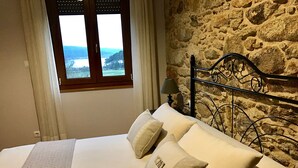 A bedroom with a stone wall, a bed with white bedding, and a window with a view of a landscape. - House in a natural setting, 200m from the beach in Costa da Morte (Balarés)