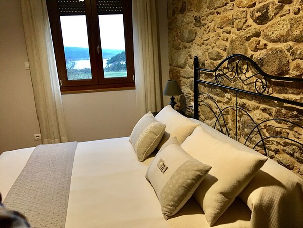 A bedroom with a stone wall, a bed with white bedding, and a window with a view of a landscape.