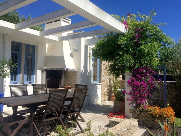 An outdoor patio with a stone table and chairs, a built-in grill, and a pergola with flowering plants.