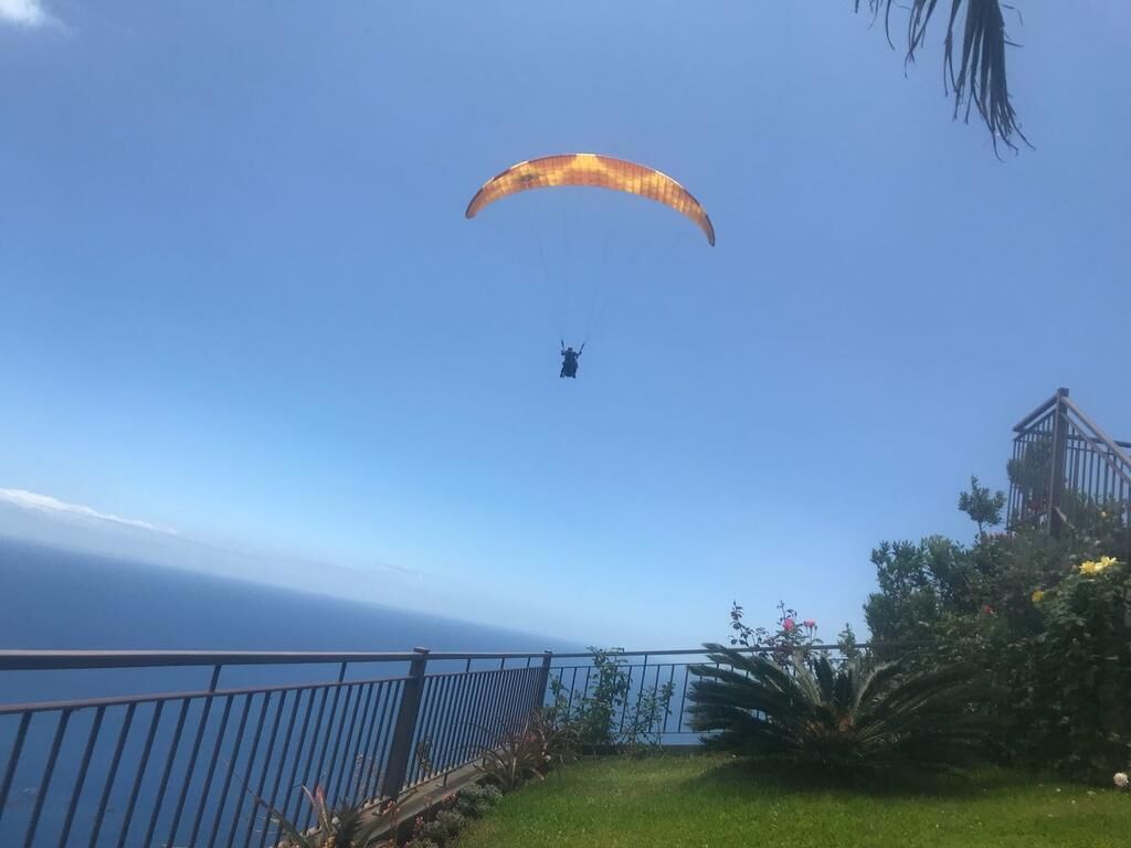 A person paragliding with a parachute above them, overlooking a coastal area with a railing and greenery.
