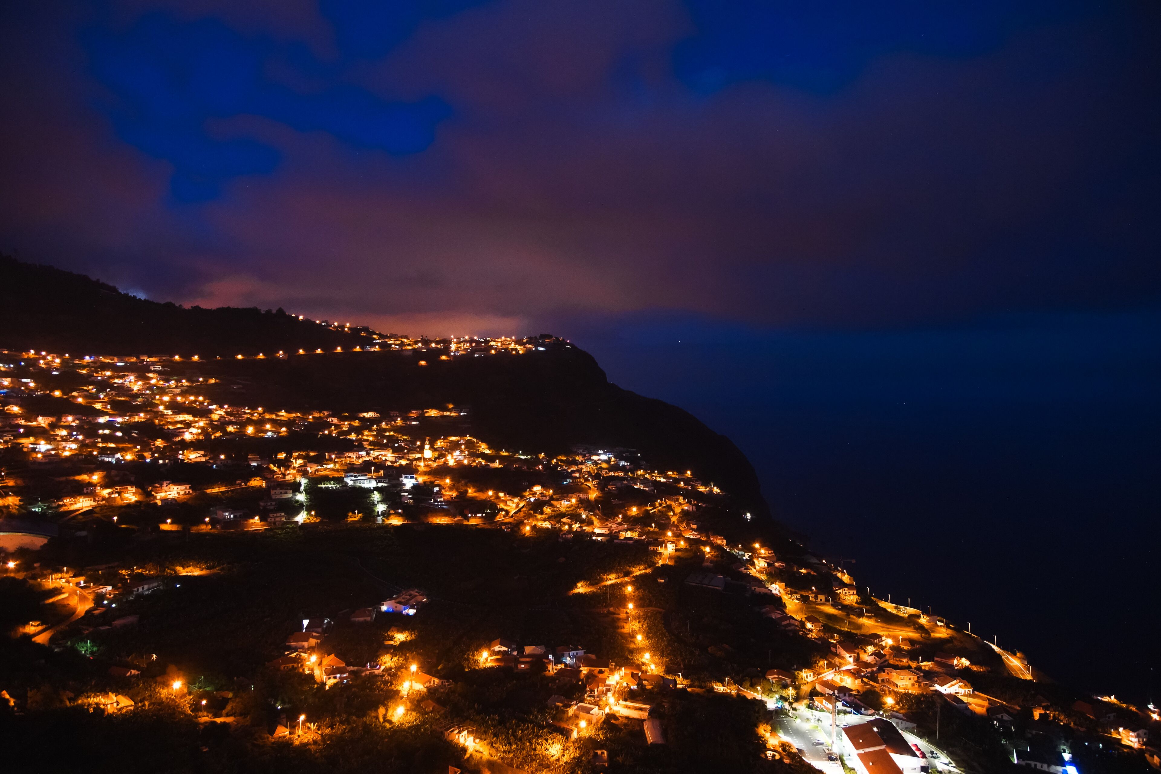 A nighttime view of a town with streetlights illuminating the area, a prominent hill in the background, and a clear sky.