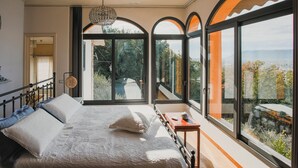 A bedroom with a large bed, a small table, and multiple windows offering a view of the outdoors.