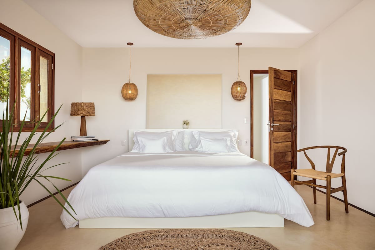 A bedroom with a large bed, a wooden chair, a potted plant, and a hanging light fixture.