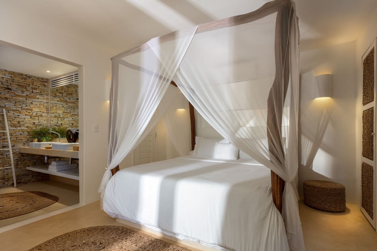 A bedroom with a canopy bed, a stone wall, a wooden ladder, a round wicker ottoman, and a white bedside table with a plant.