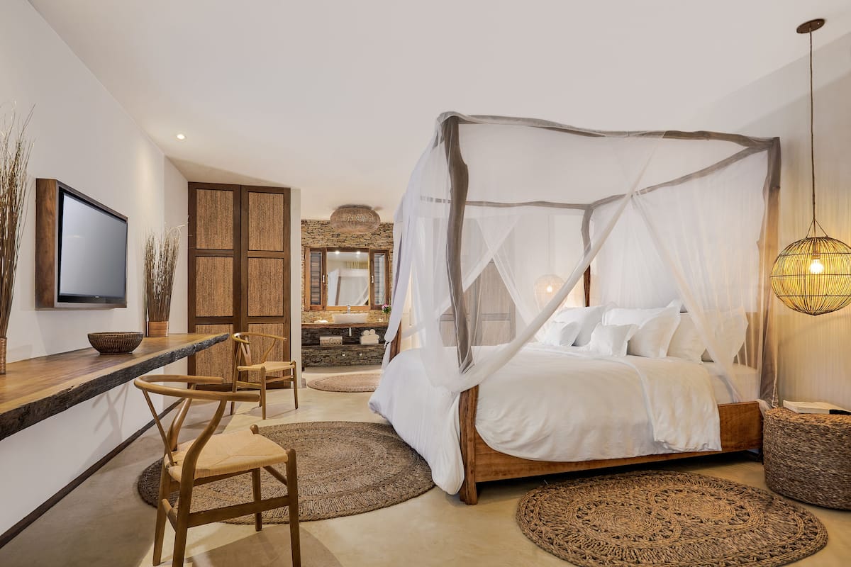 A bedroom with a four-poster bed, a bedside table, a TV, and a bathroom with a stone sink and mirror.