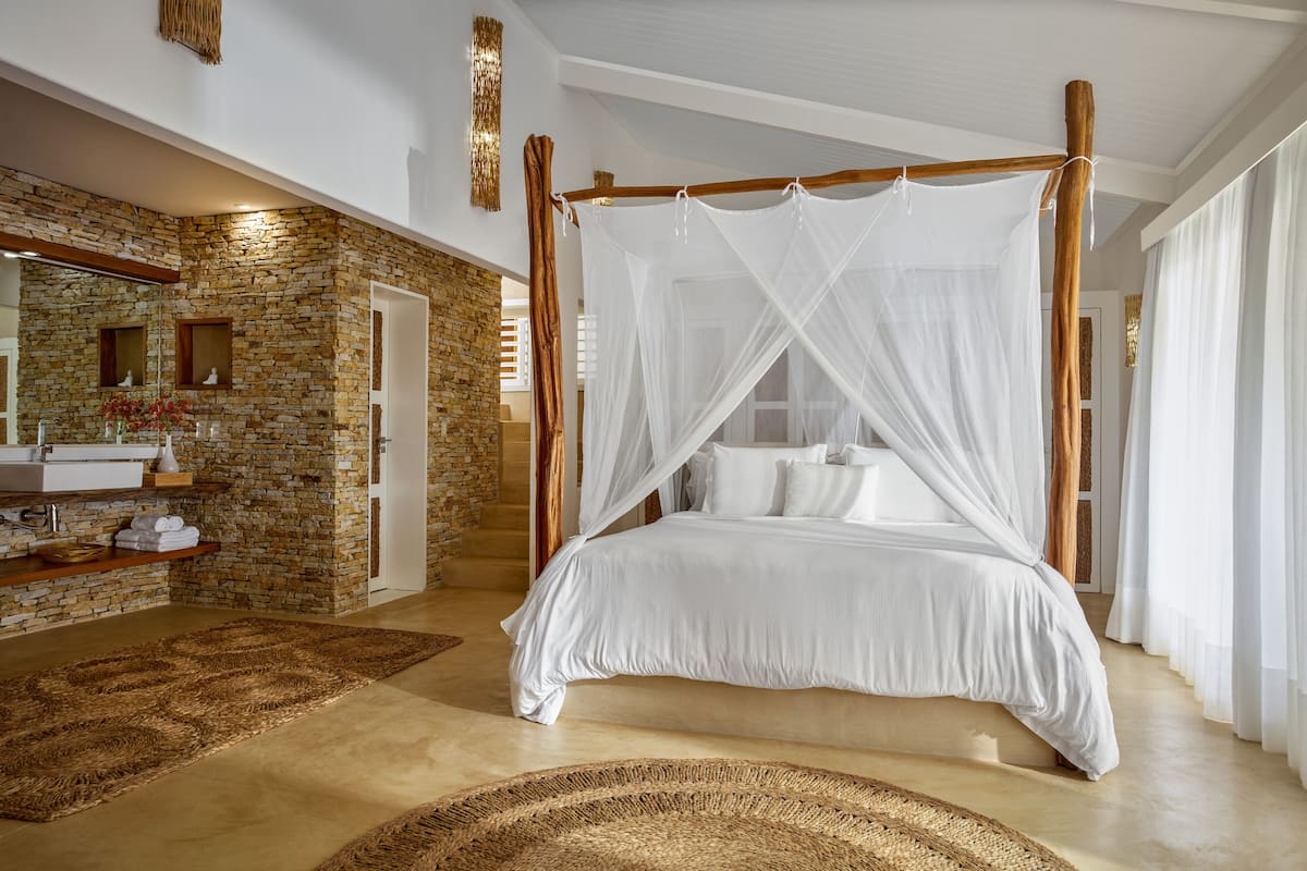 A spacious bedroom with a large bed, a stone accent wall, a wooden canopy bed, and a modern bathroom with a stone backsplash.