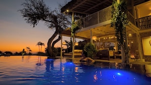 A poolside area with a large tree, a covered seating area, and a building with a balcony.