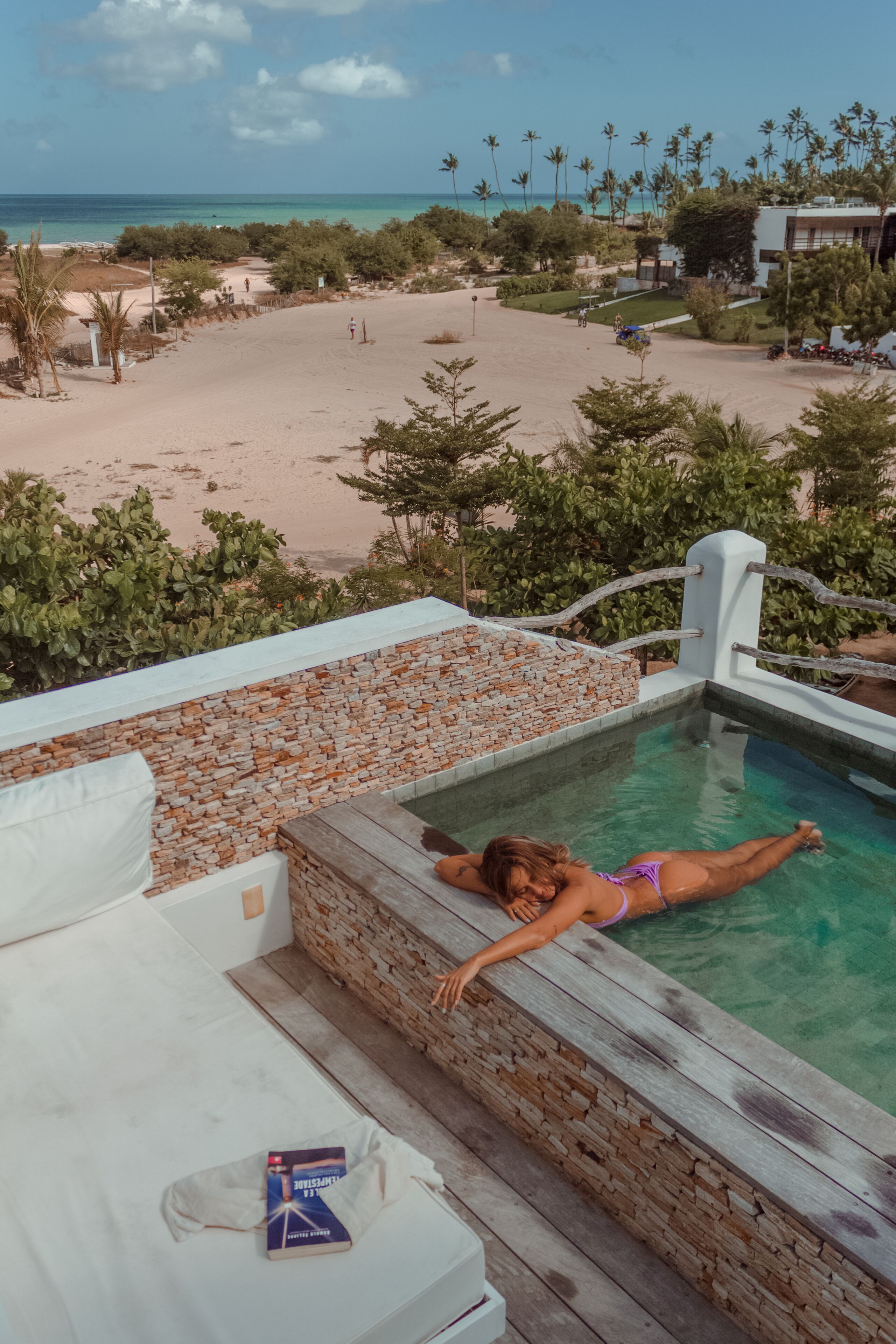 A pool area with a person relaxing, surrounded by a beach view and palm trees.