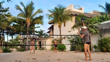 Jogo de voleibol de praia numa quadra de areia, rodeado por palmeiras e um edifício com telhado de palha.