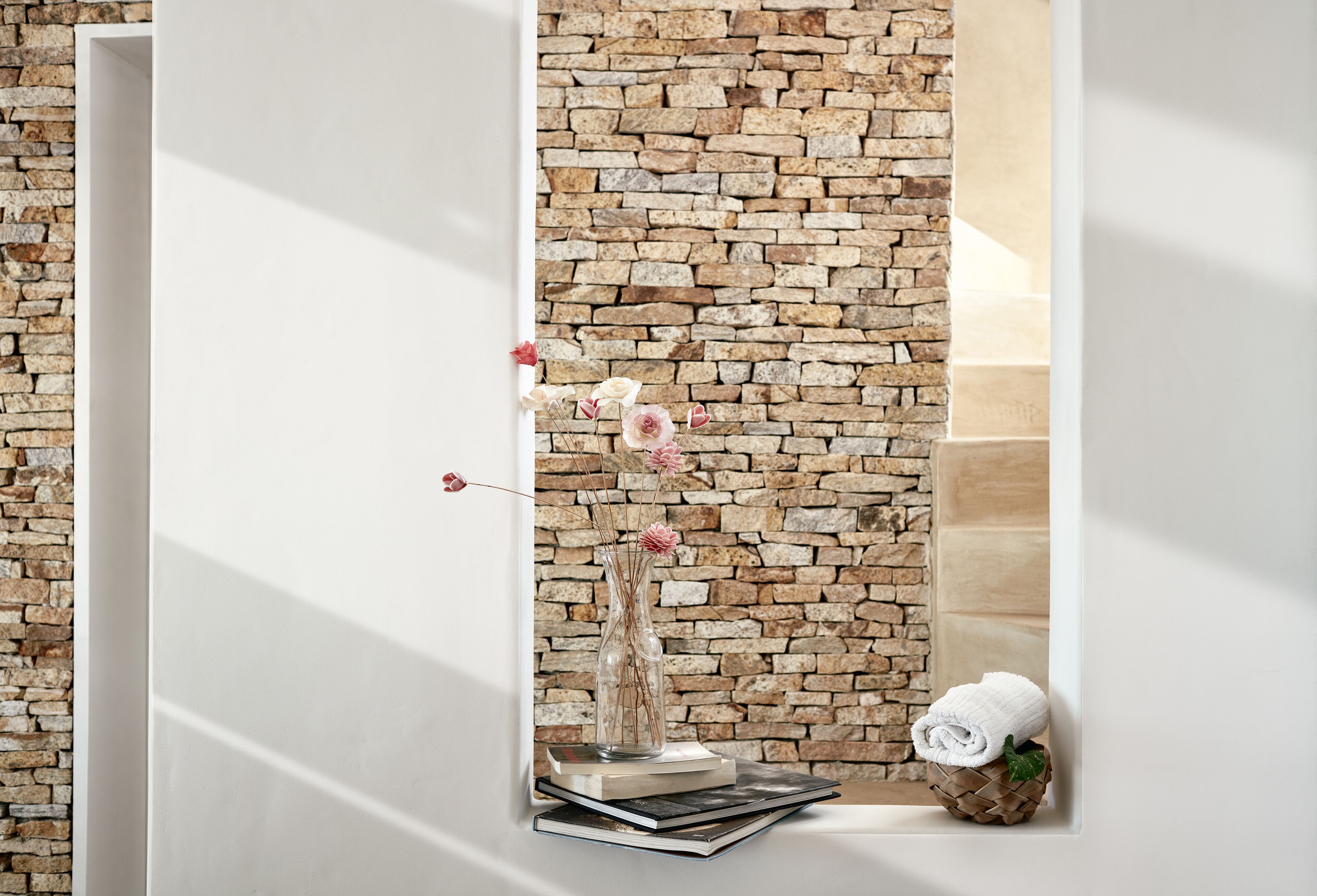 A room with a brick wall, a white door, a vase with flowers, books, and a rolled towel on a wooden surface.