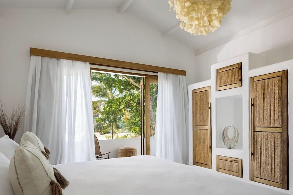 A bedroom with a large bed, white curtains, a wooden wardrobe, and a view of greenery outside.