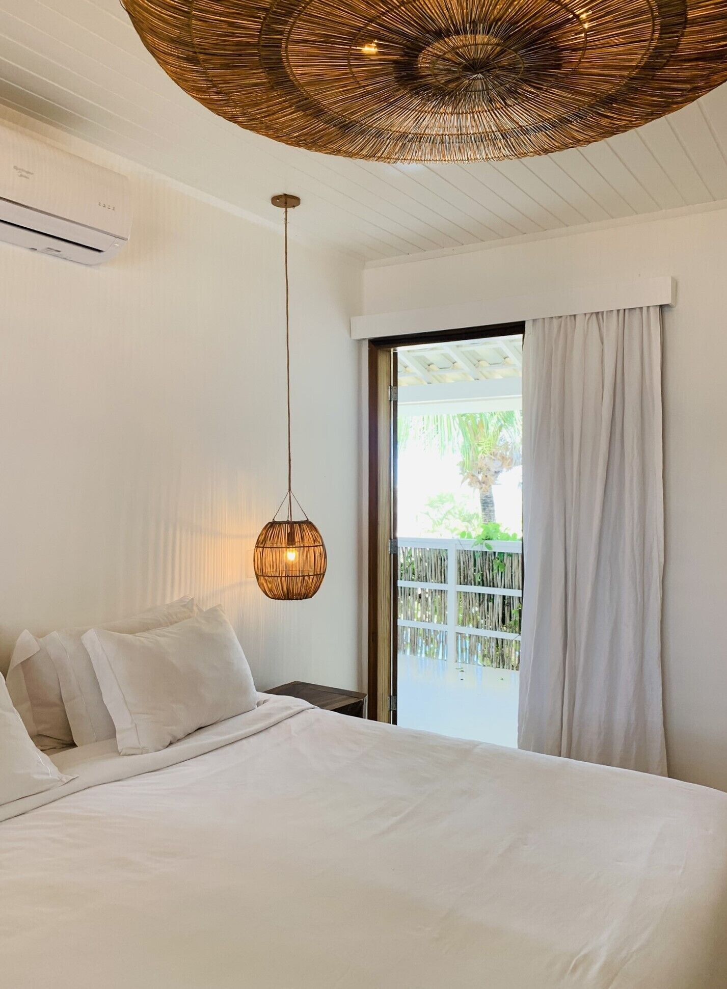 Een slaapkamer met een bed, een hangende rotanlamp, een raam met uitzicht op groen en een plafondventilator.