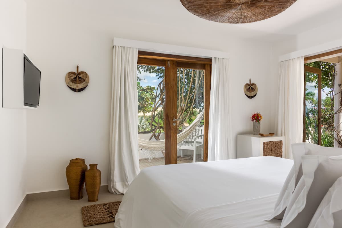 A bedroom with a large bed, white curtains, a TV, and a view of trees through the window.