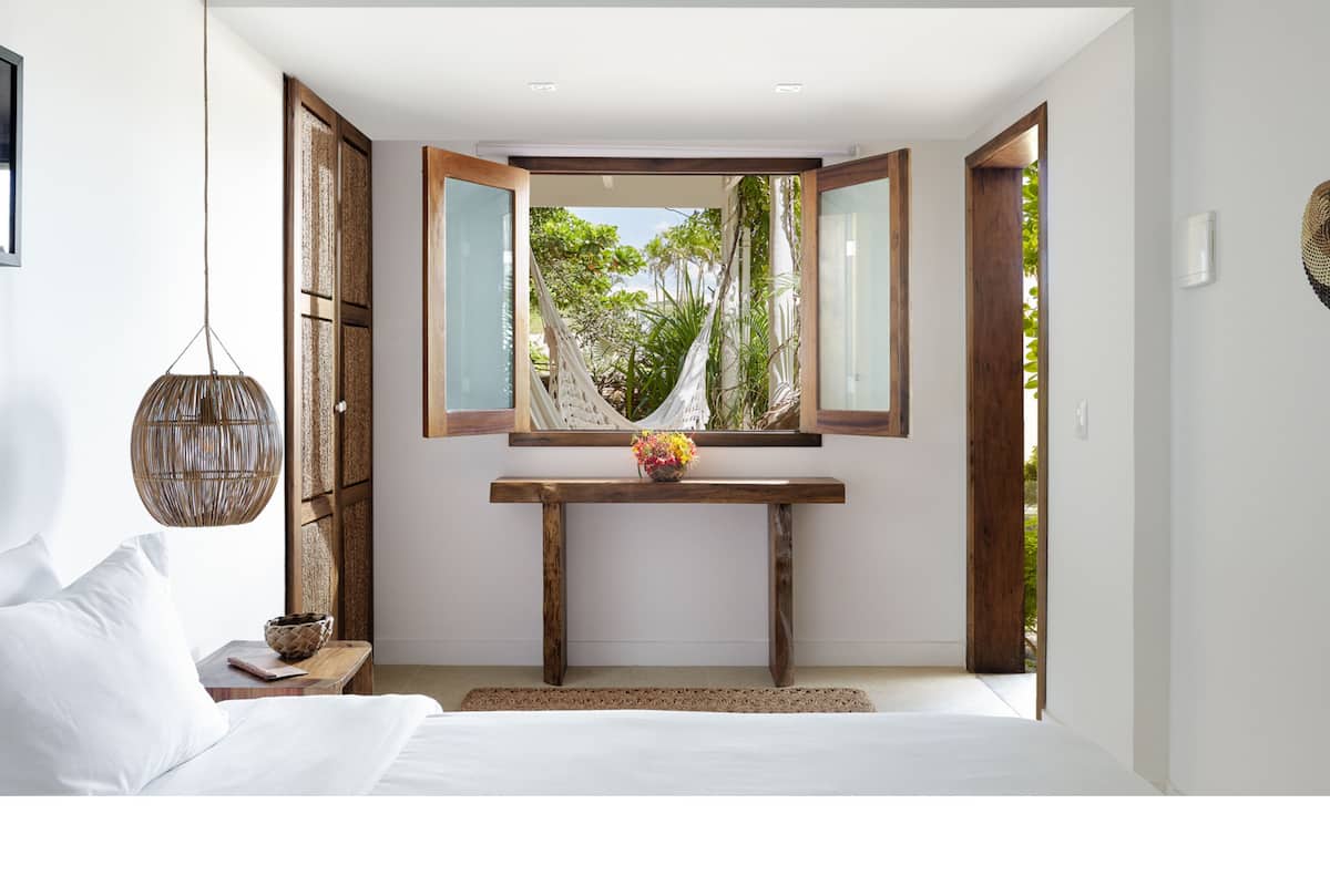A bedroom with a bed, a bedside table with a lamp, a console table with a vase, and a view of greenery through a window.