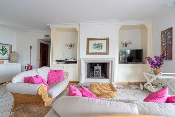 Un salon avec un canapé blanc, un fauteuil en osier, une cheminée, un téléviseur et un meuble blanc.