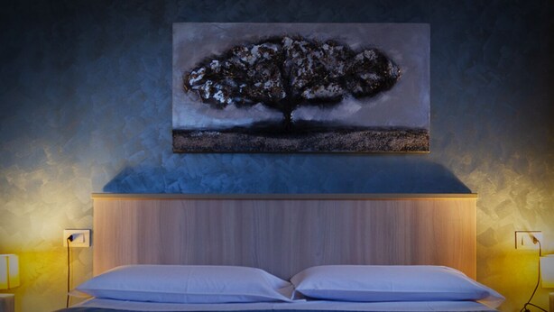 A bedroom with a bed, two bedside lamps, a wooden headboard, and a painting of a tree on the wall. - The house of Bice - accommodation 2 (Siracusa)
