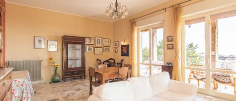 Una habitació amb un sofà blanc, una taula de dinar amb cadires, una làmpada penjant i una finestra gran amb cortines.
