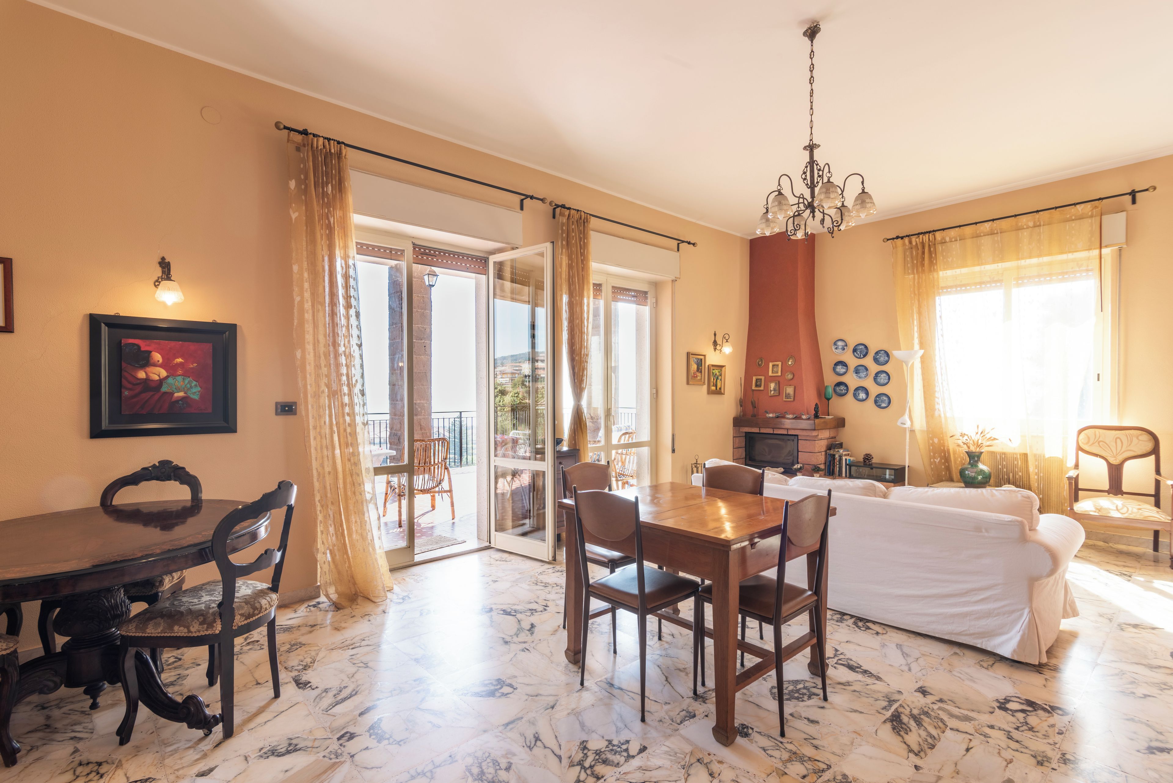 A dining area with a marble floor, a wooden table, and chairs. There is a white sofa and a chandelier in the room.