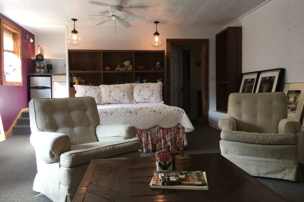 Una sala de estar acogedora con una cama, dos sillones, una mesita de centro con libros, un ventilador de techo y una estantería llena de objetos.