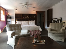Uma sala de estar acolhedora com uma cama, duas poltronas, uma mesa de centro com livros, um ventilador de teto e uma estante repleta de objetos.