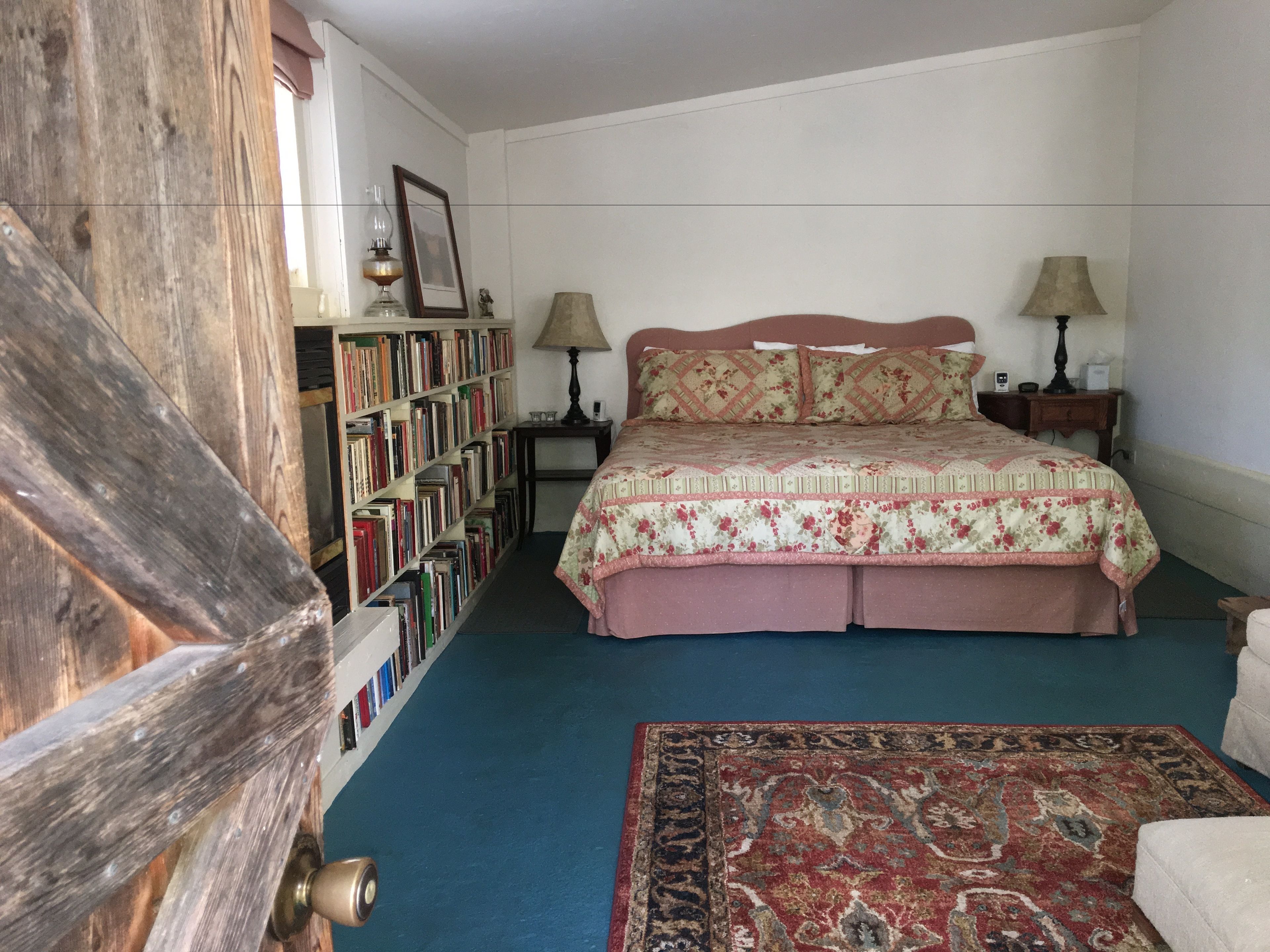 A bedroom with a wooden door, a bookshelf, a bed with floral bedding, two bedside lamps, and a patterned rug.