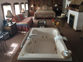 Um quarto espaçoso com uma cama grande, um espaço de estar com uma poltrona e uma mesa, uma banheira e piso de madeira.
