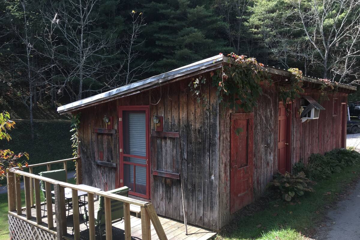 Una cabaña rústica de madera con puerta roja, un pequeño porche y una valla de madera.