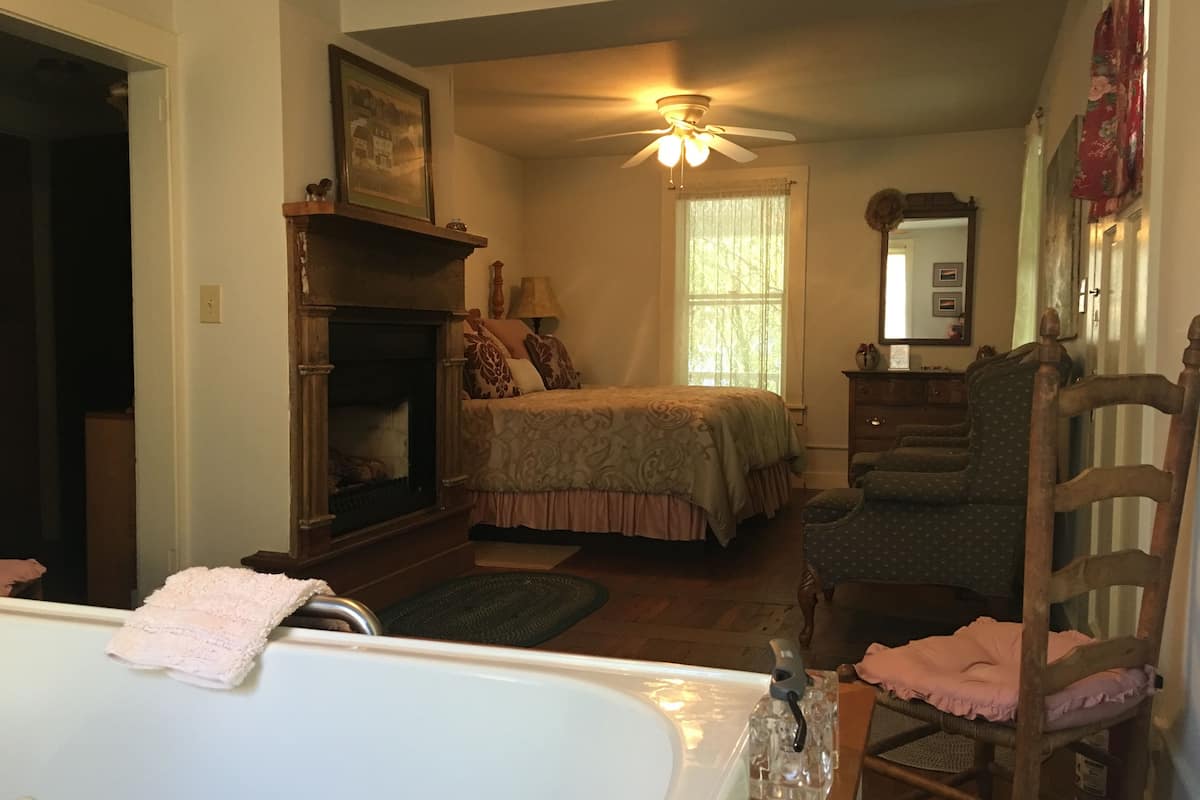 Un dormitorio con cama, una silla, chimenea, ventilador de techo y ventana.