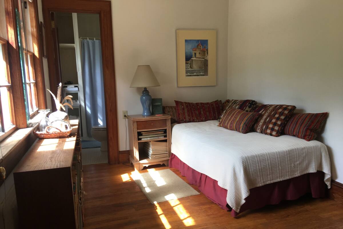 Un dormitorio con suelo de madera, una cama con edredón blanco y varias almohadas, una mesita de noche con lámpara, un cuadro enmarcado en la pared y una ventana por donde entra luz solar.