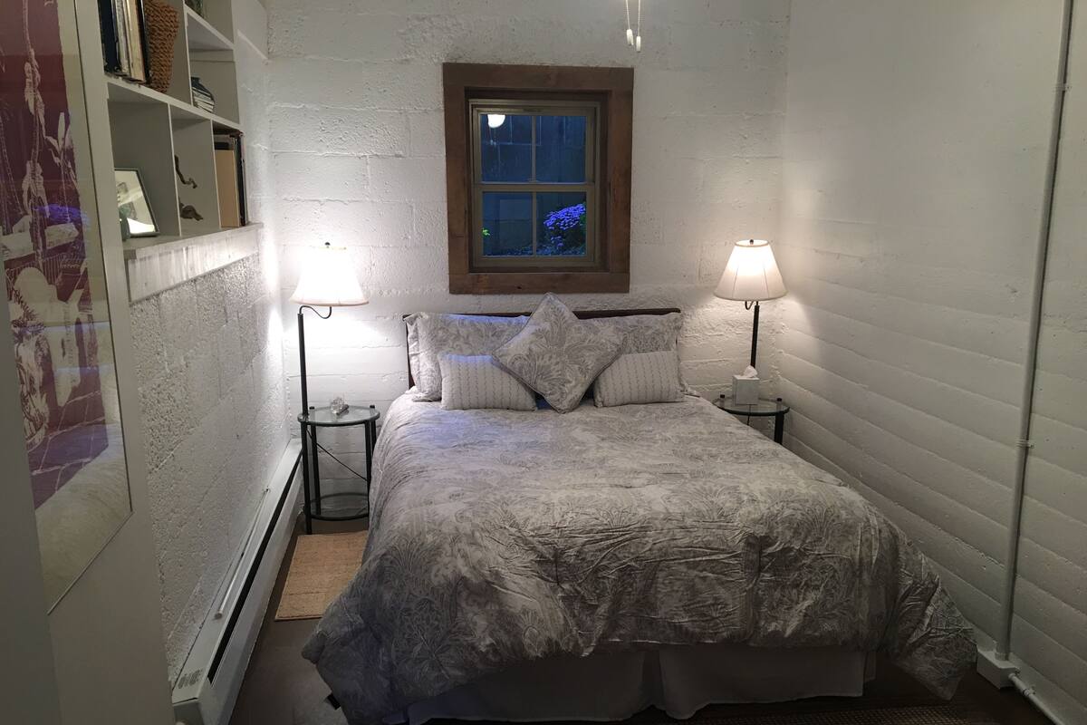 Un dormitorio pequeño y acogedor con cama, dos lámparas de noche, una mesita de noche, una ventana y una estantería.