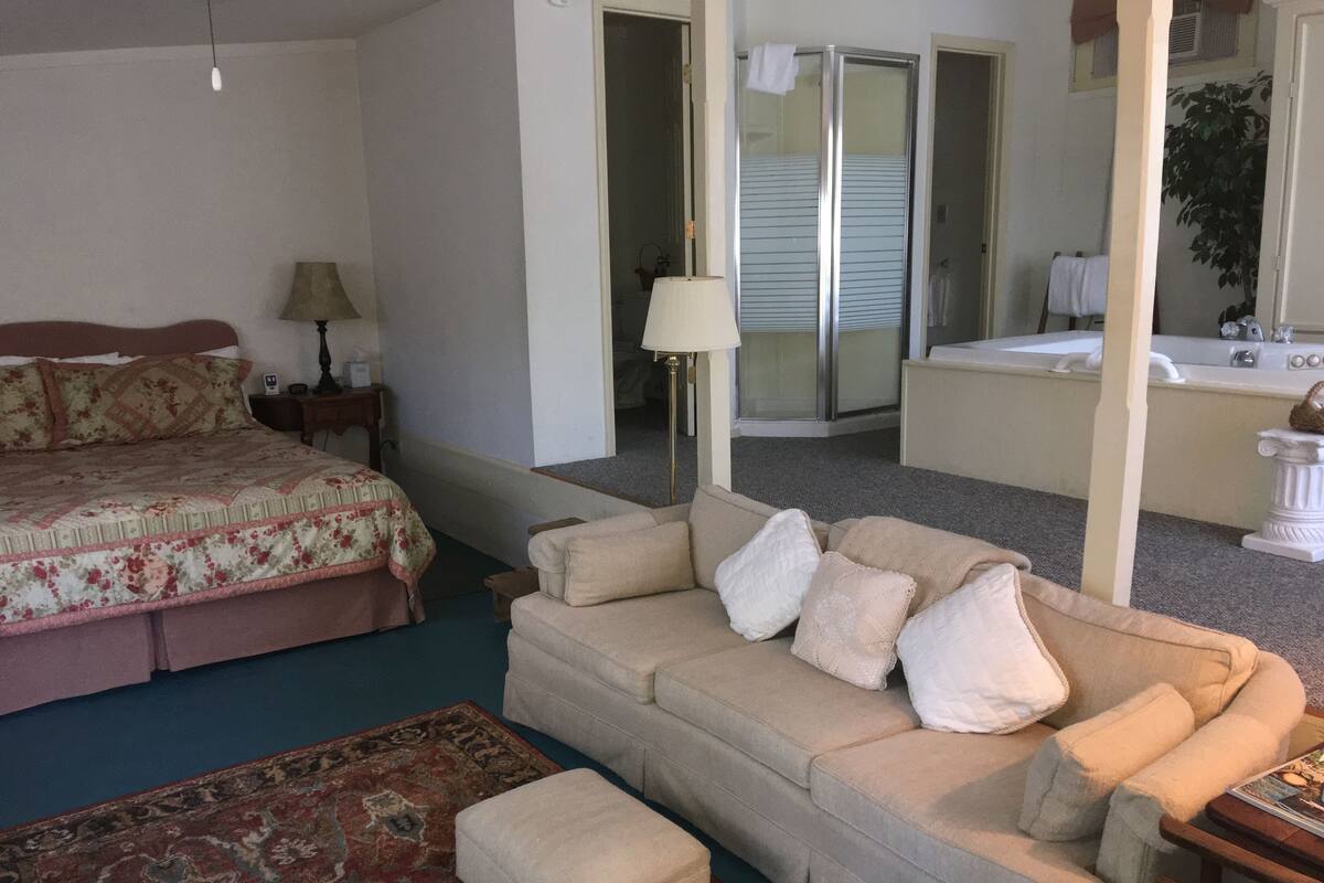 Un dormitorio con una cama, un sofá, una mesita, una lámpara y un baño visible a través de una puerta abierta.